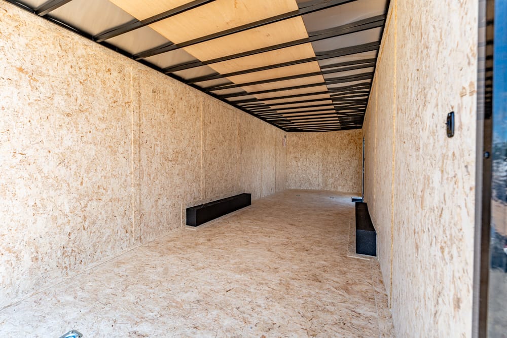 Interior of an empty plywood cargo trailer with tan walls and ceiling beams, and a black storage box along the left wall.