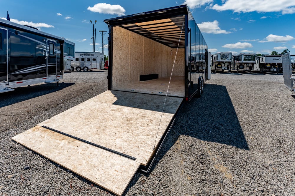 Open cargo trailer with ramp down, showing plywood interior on a sunny gravel lot with other trailers nearby