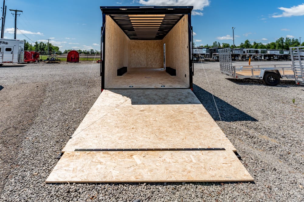 Open cargo trailer with a wooden ramp extended to the ground, ready for loading on a gravel lot