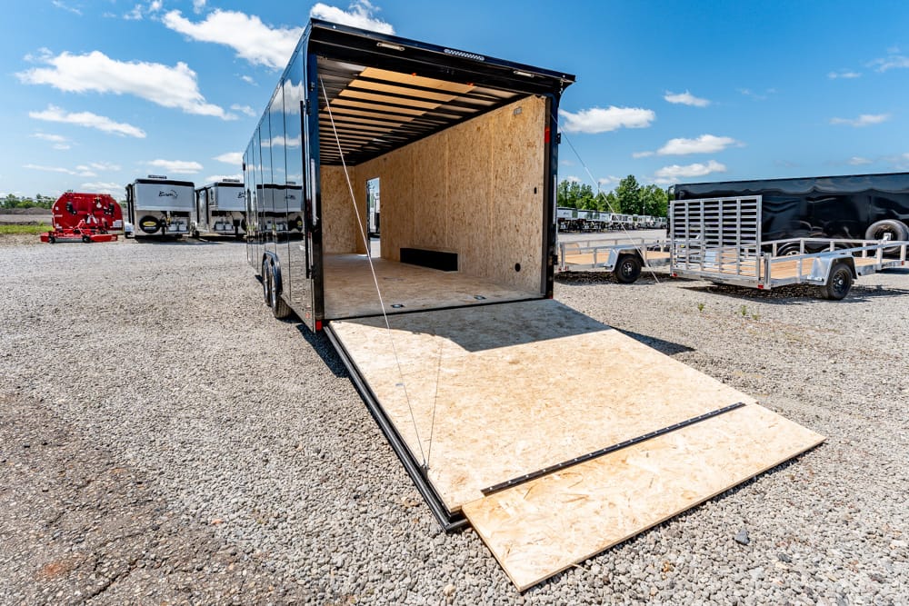 Open black enclosed trailer with ramp lowered, showing plywood interior ready for loading in a gravel lot with other trailers nearby on a sunny day.