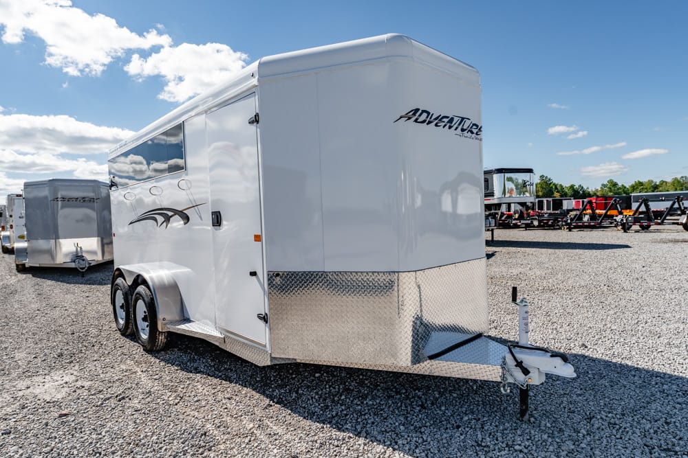 White enclosed cargo trailer (dually axles) with diamond-plate front, parked on gravel, other trailers in the background under a blue sky.