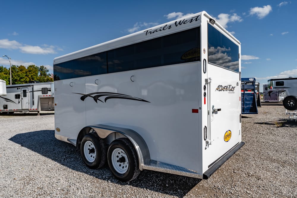 White Trails West horse trailer with black decorative stripe, parked on a gravel lot under a blue sky with other trailers nearby