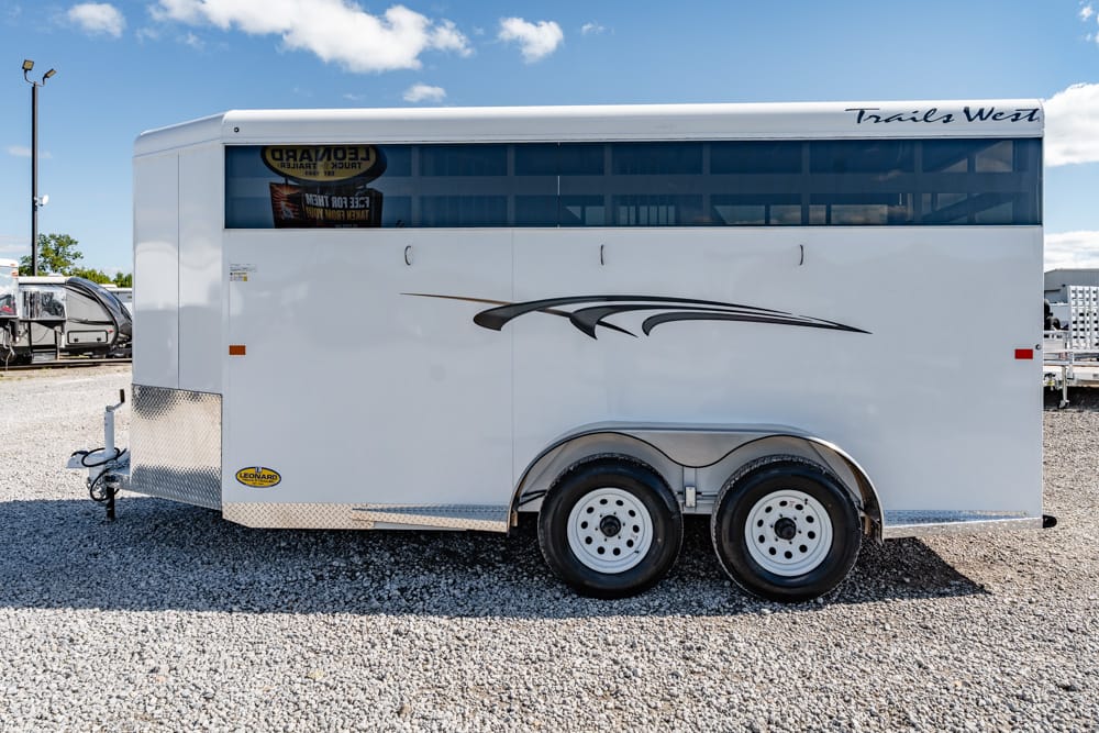 White enclosed horse trailer with dual axles and decorative black swoosh graphics on a gravel lot under a blue sky, hitch visible at the front.