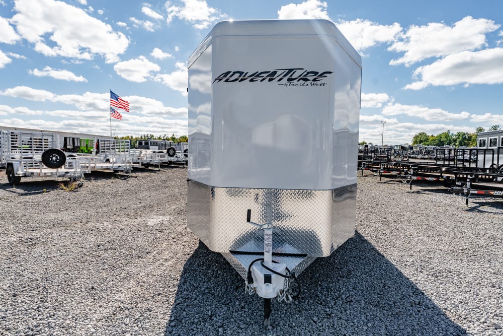 Front view of a white Adventure enclosed trailer with diamond-plate nose on a gravel lot, hitch in foreground and American flag in the background.