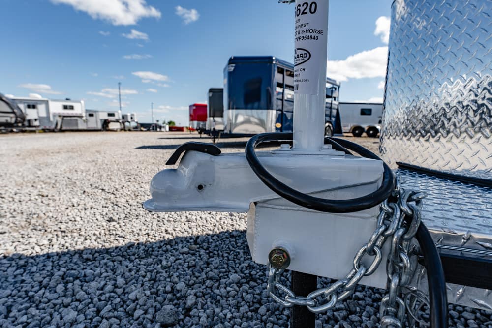 Close-up of a trailer hitch with a safety chain and jack on a gravel lot, RVs in the background under a blue sky.