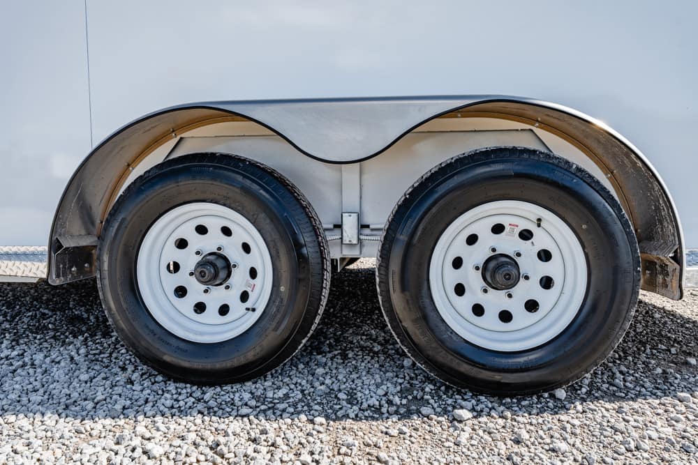 Twin trailer wheels with white rims and black tires under a curved silver fender on gravel ground.