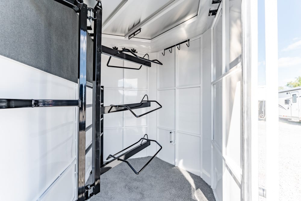 Interior of a white horse trailer with black metal saddle racks and wall hooks, carpeted floor visible.