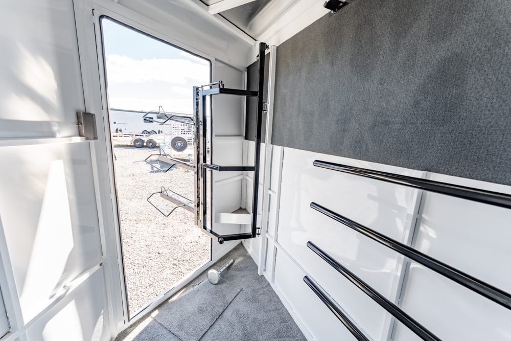 Interior of a white trailer's tack room with a pull-out cabinet and chrome rails, door open to a gravel lot outside.