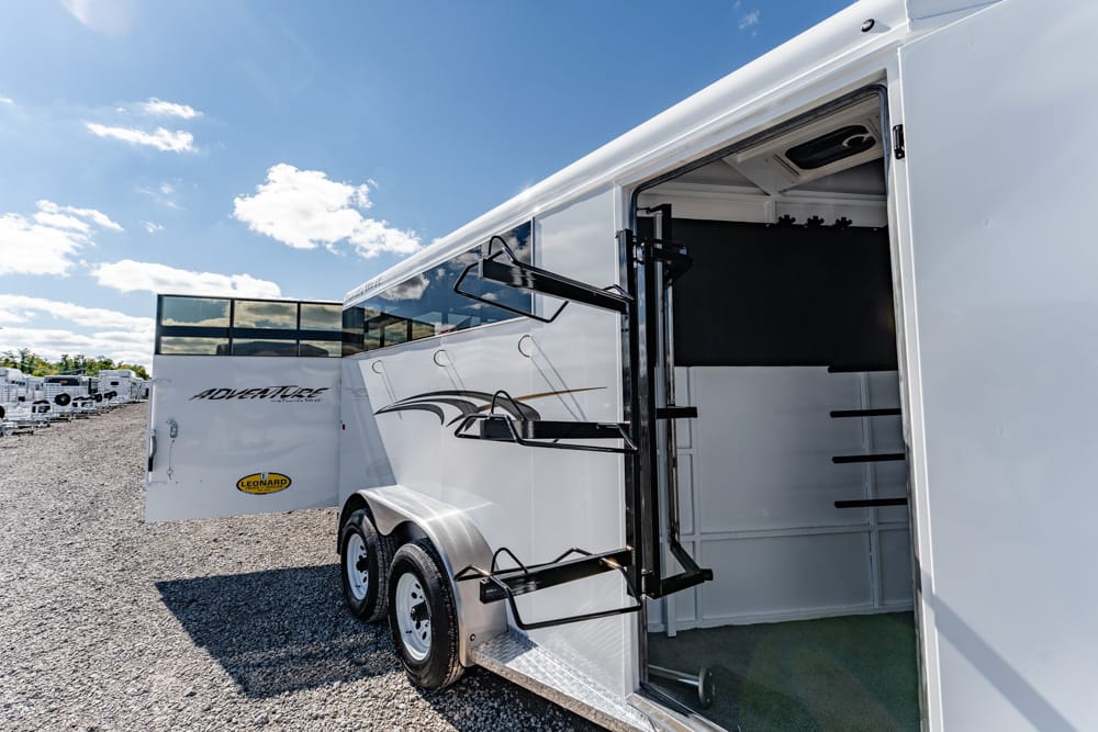 White horse trailer with side door open, revealing interior tack storage racks on a gravel lot under a blue sky.