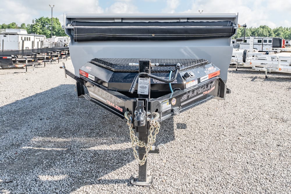 Front view of a Horizon Trailers dump trailer parked on a gravel lot, showing the hitch, safety chains, and toolbox in the foreground.