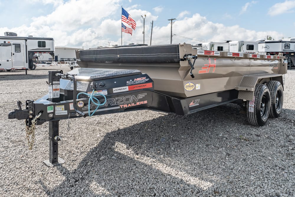 Large beige boat trailer with dual axles on a gravel lot, secured with a cover and towing hitch visible; American flag in the background.