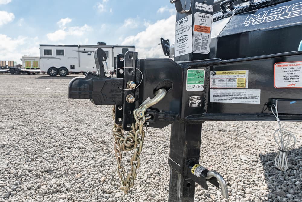 Close-up of a trailer hitch with safety chains and a black coupler, on a metal frame, in a gravel lot with RVs in the background.