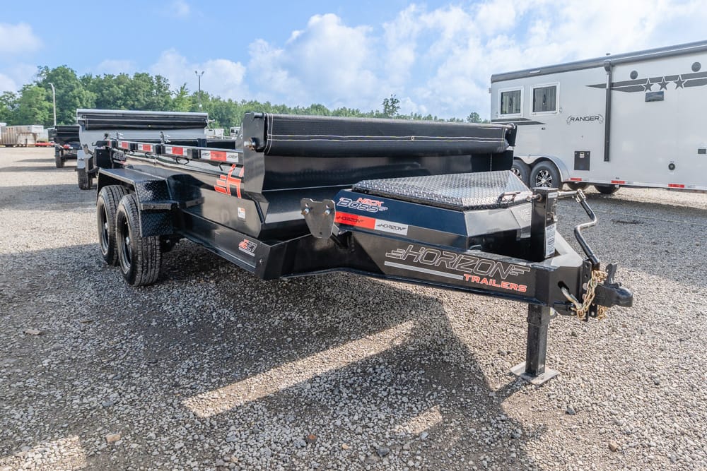 Black Horizon dump trailer with dual axles on a gravel lot, hitched to a vehicle, with an RV in the background (industrial trailer in a dealership yard).