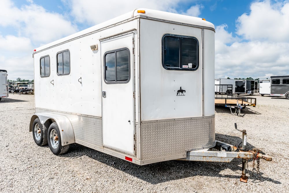 White horse trailer on a gravel lot with dual-axle wheels and a front hitch area.