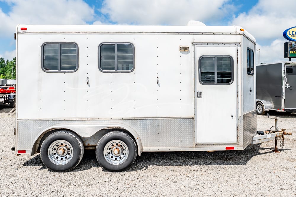 White utility trailer with two windows and a side door, sitting on gravel with a diamond-plate lower panel.