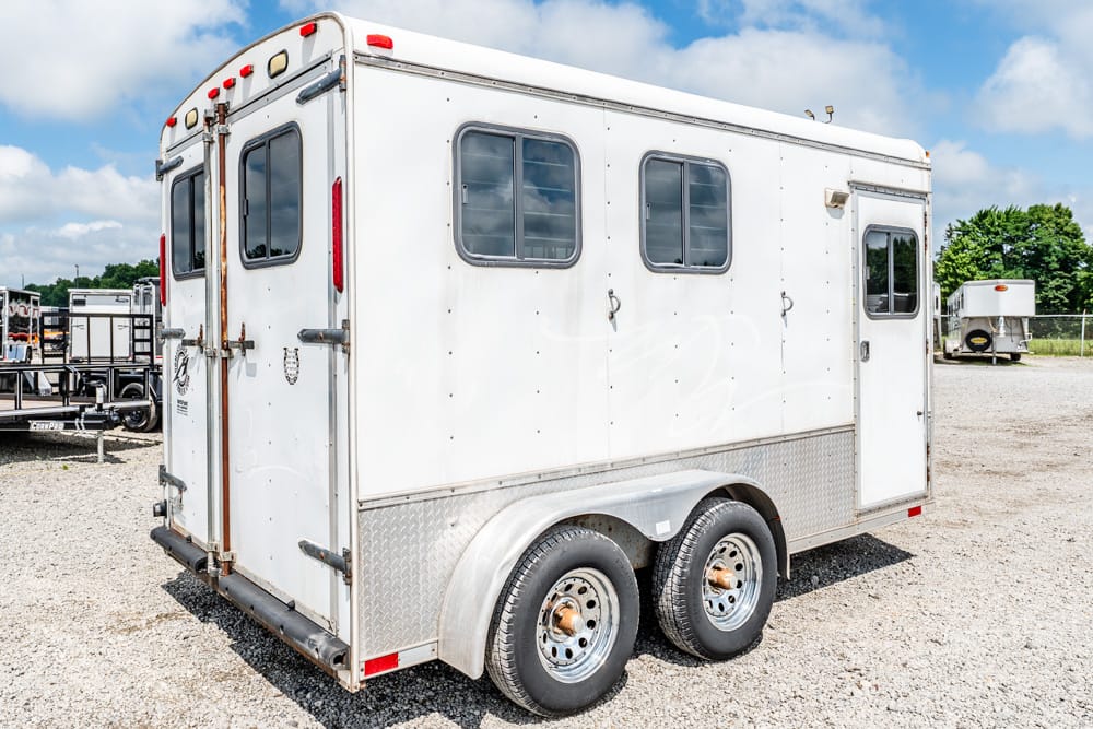 White horse trailer with two axles and side windows parked on a gravel lot under a blue sky.