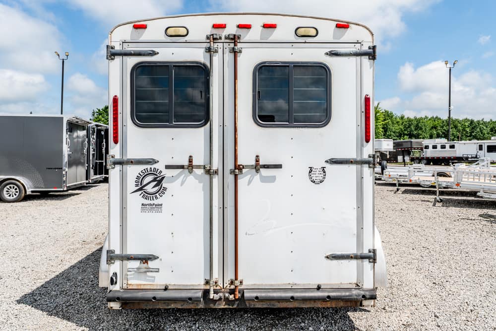 Back view of a white horse trailer with two small rectangular windows and heavy metal latches, parked on a gravel lot with other trailers nearby