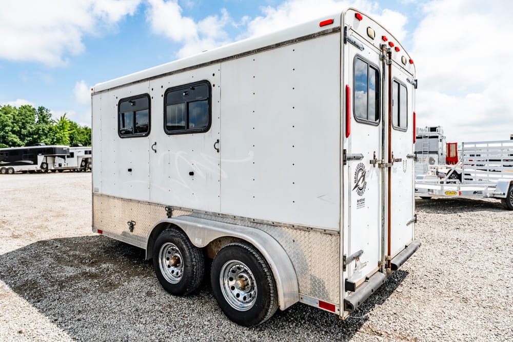 White enclosed dual-axle trailer with two small side windows and rear double doors on a gravel lot.
