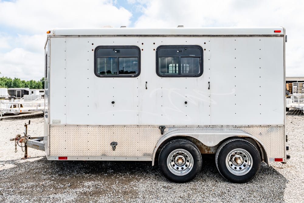 White two-window travel trailer with diamond-plate lower siding and tandem wheels on a gravel lot.