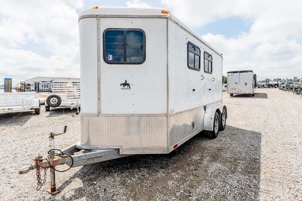 White horse trailer with two side windows and a diamond-plate lower front, parked on a gravel lot.