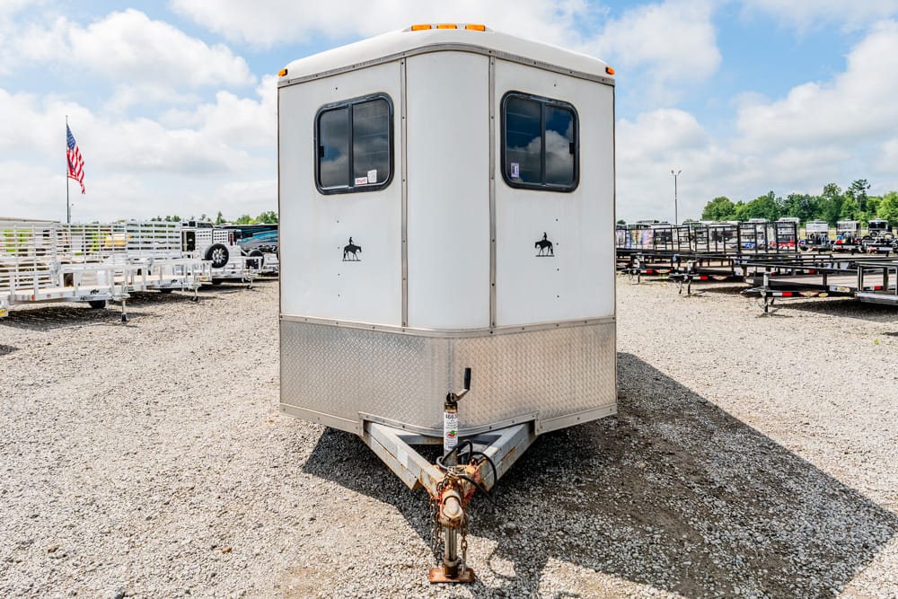 White horse trailer with two small horse decals and a diamond-plate front, parked on a gravel lot on a sunny day (front view).