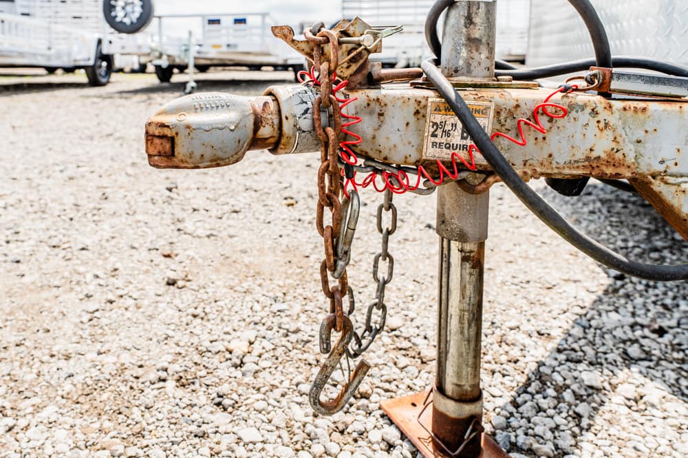 Rusted trailer hitch with chains hanging from a metal jack on gravel in a storage lot.