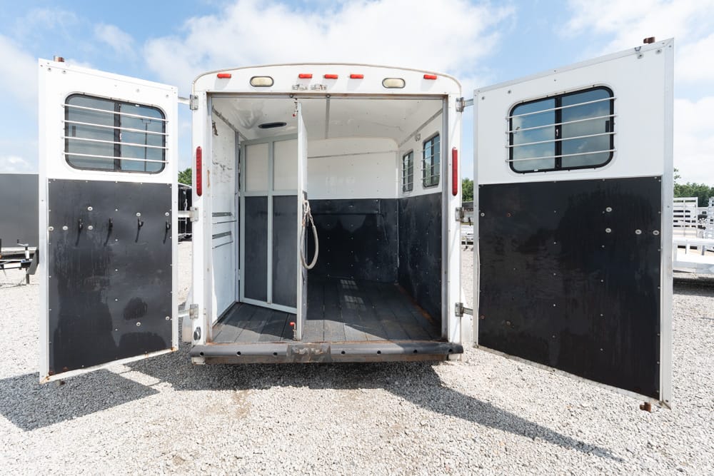 Open horse trailer with rear doors wide, showing an empty interior and partition on a gravel surface under a blue sky.