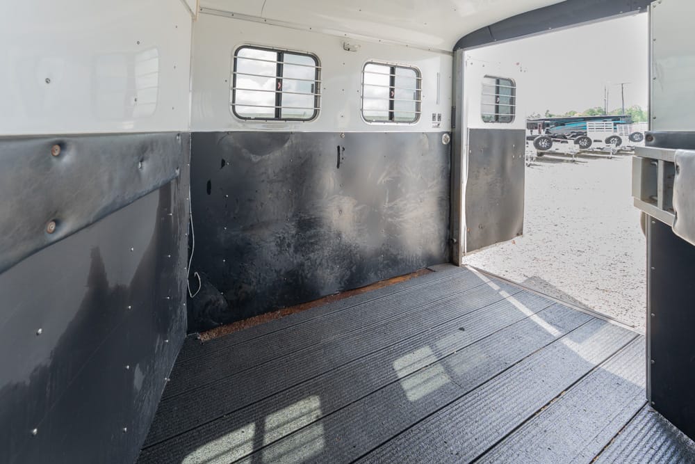 Interior of a white horse trailer with black lower walls, gray non-slip floor, and open side door; outside show trailers in a lot.