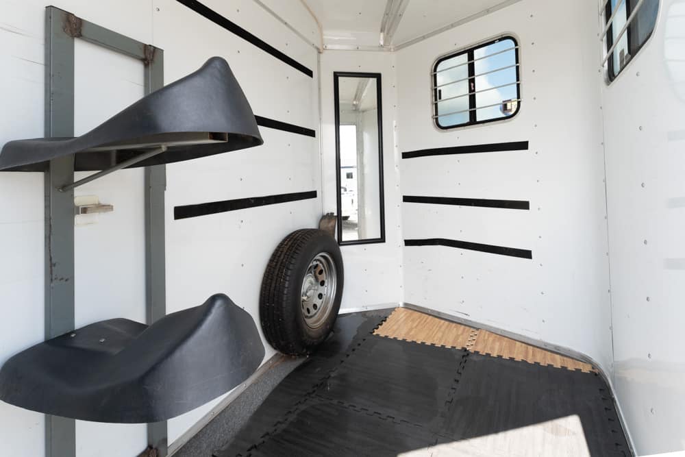 Interior of a white horse trailer with saddle racks, a spare tire, and padded flooring mats for loading animals or gear.