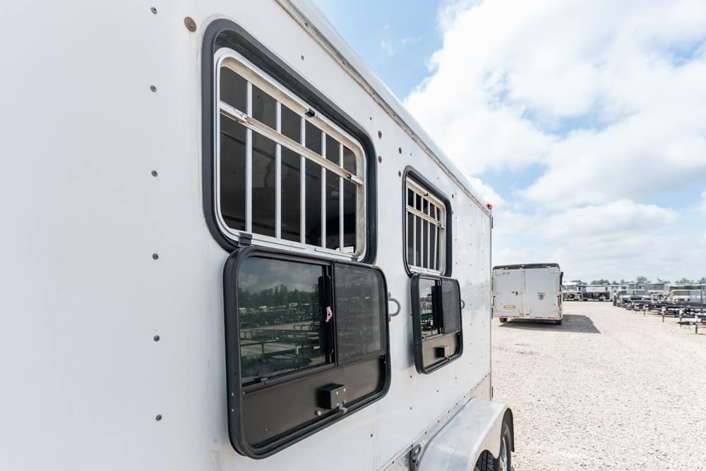 White horse trailer with barred windows, parked on a gravel lot under a partly cloudy blue sky.