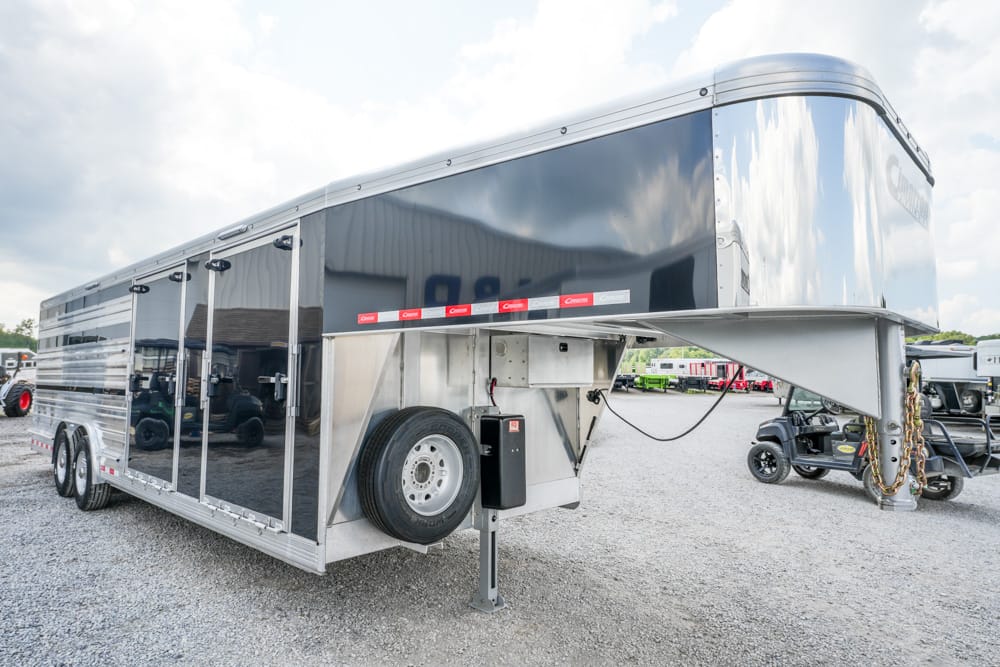 Large aluminum gooseneck cargo trailer parked on a gravel lot, with a spare tire mounted on the side and reflective panels visible underneath the trailer's overhang.
