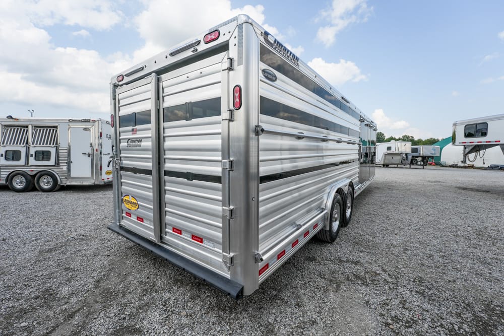 Silver aluminum horse trailer with rear double doors in a gravel lot, other trailers visible in the background under a blue sky.