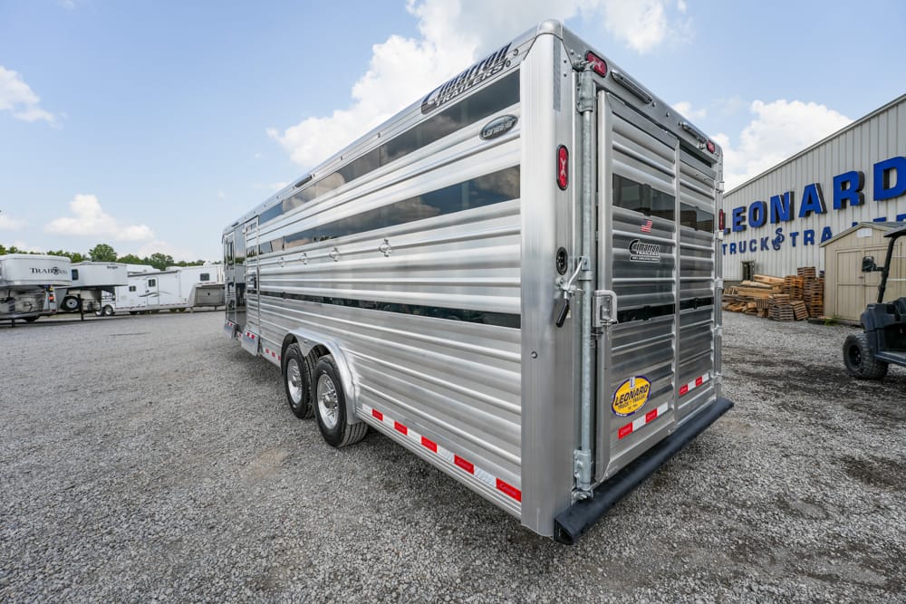 Rear view of a silver aluminum livestock trailer with dual-axle wheels, parked on a gravel lot near a building with a Leonard sign
