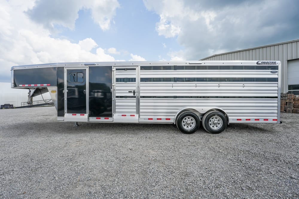 Silver aluminum horse trailer with dual-axle wheels parked on gravel, showing a black front section and gooseneck hitch at the front.