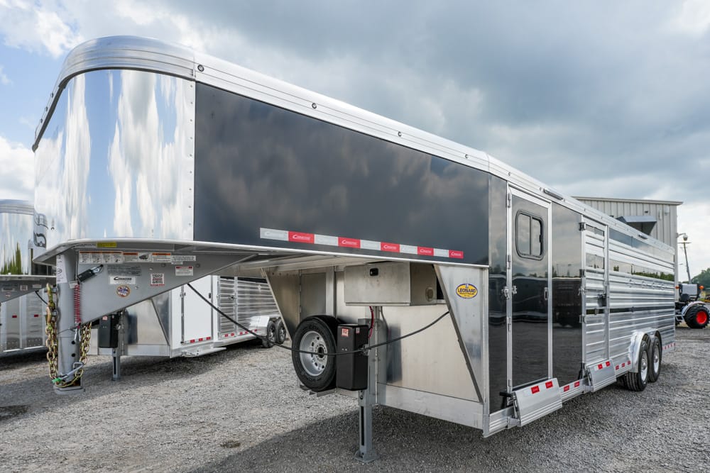Large aluminum gooseneck trailer with living quarters, parked on a gravel lot, showing dual axles and side access doors.