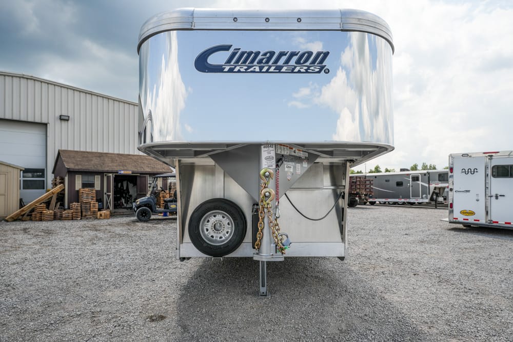 Shiny Cimarron Trailers gooseneck trailer in a gravel lot, with a spare tire, hitch, and chained coupler at the front.