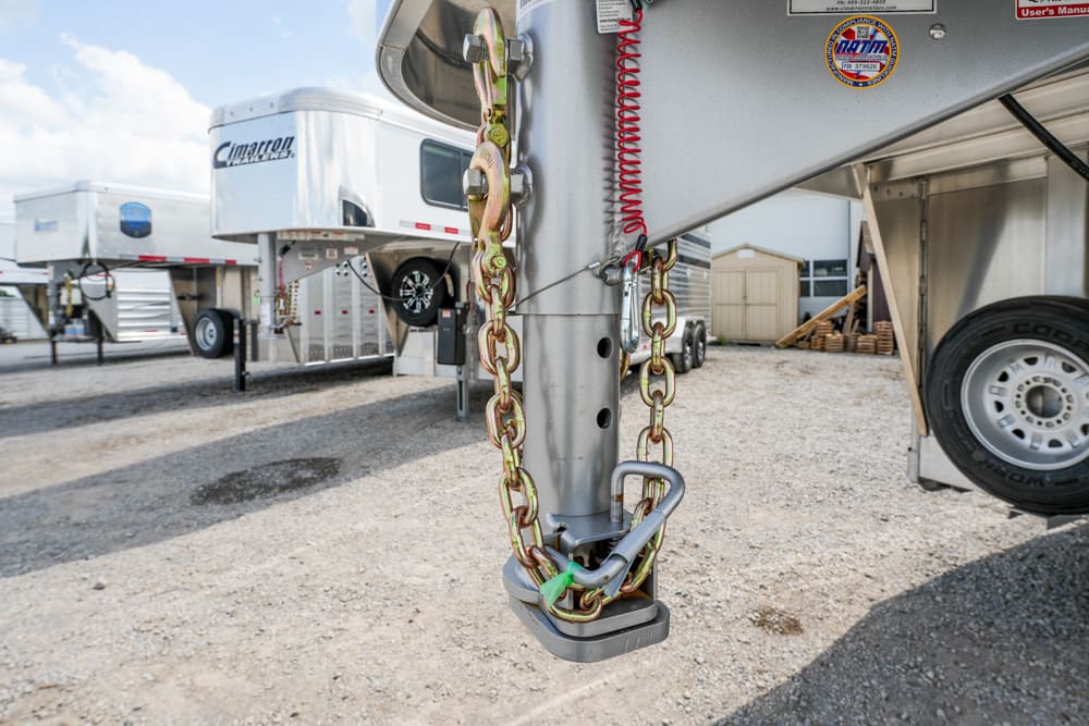 Close-up of a heavy-duty trailer hitch with safety chains and a lock, attached to a gooseneck trailer in a gravel lot with other trailers in the background.