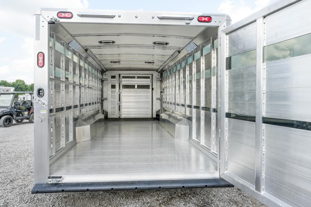 Interior of an empty aluminum livestock trailer with side stalls and a rear ramp open, bright daylight visible outside.