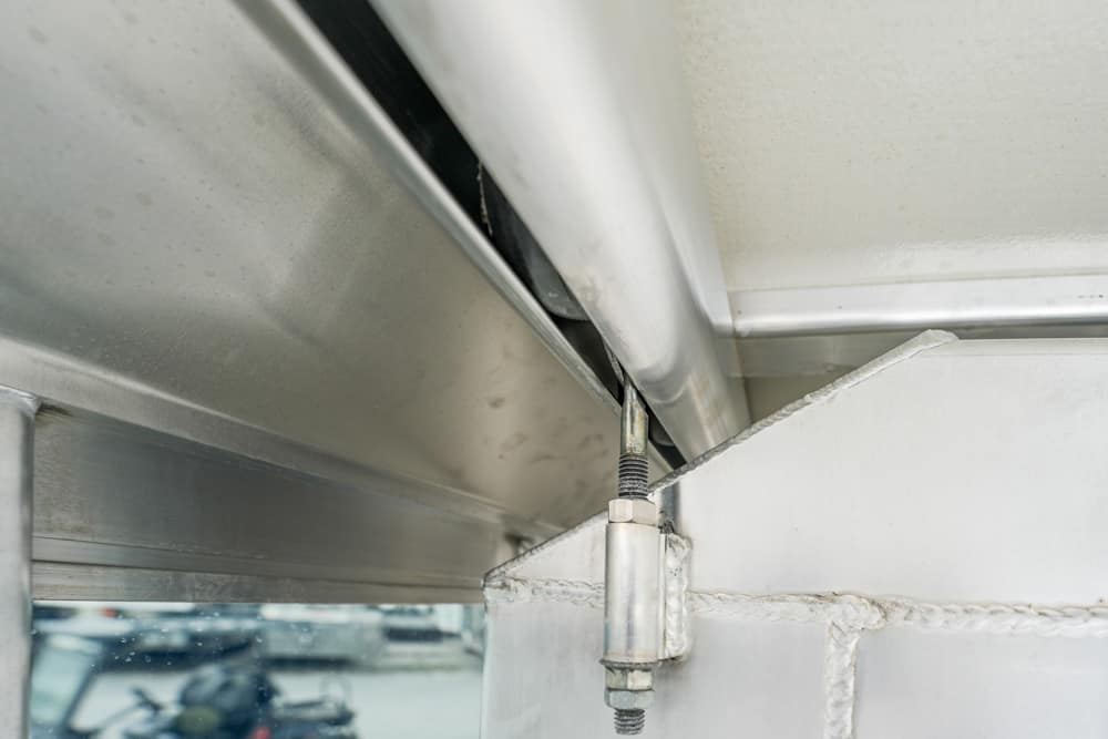 Close-up of a metal hinge with a coiled spring and bolts securing a beam, part of an outdoor frame or awning.