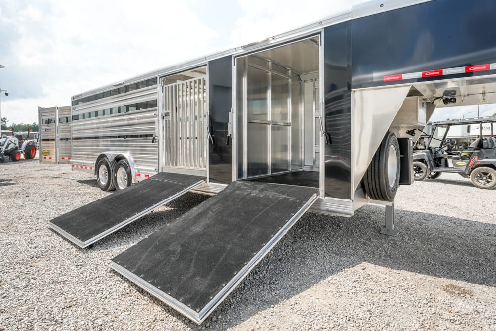 Aluminum livestock trailer with two loading ramps extended, doors open, on a gravel lot at a truck yard.
