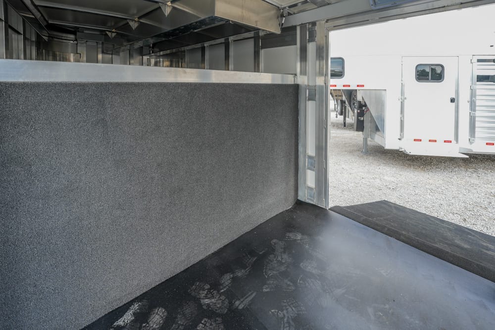 Interior of a horse trailer with gray padded wall and textured rubber floor, open doorway reveals a white trailer outside on gravel outside.