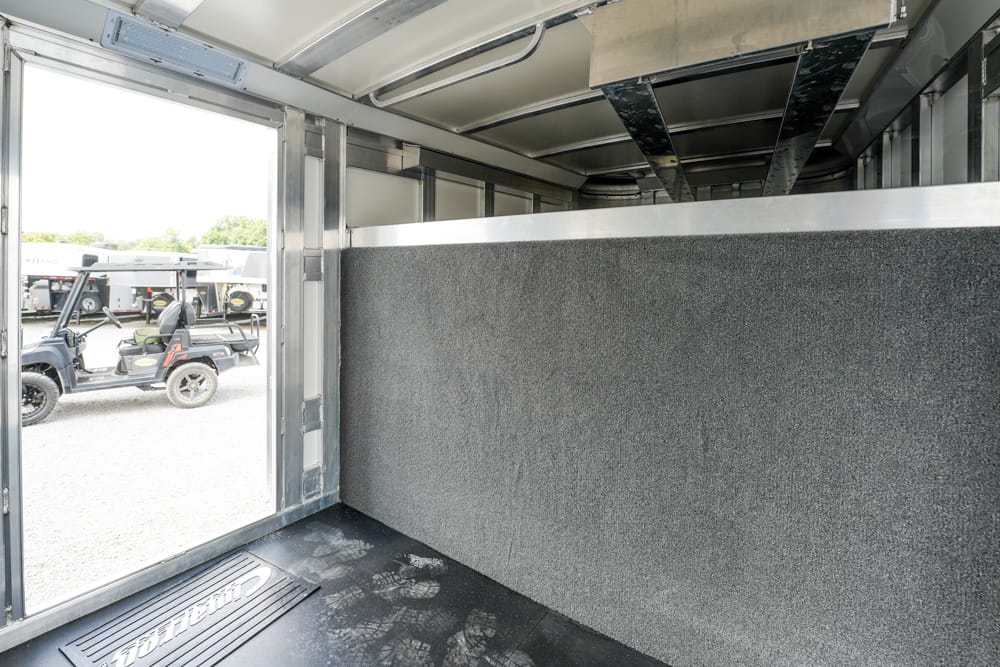 Interior of an empty metal cargo trailer with gray padded wall, dark rubber floor, and exposed ceiling beams; open rear door reveals parked golf carts outside.