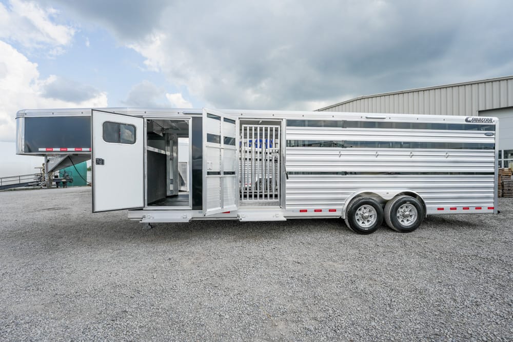 Large aluminum two-axle horse trailer on a gravel lot, side and rear doors open revealing horse stalls and interior compartments under a cloudy sky.