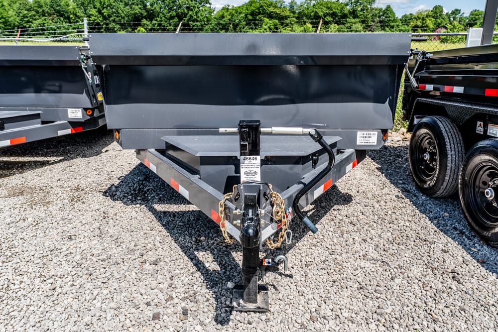 Front view of a gray dump trailer hitch with safety chains and crank, parked on gravel next to other trailers in a lot.
