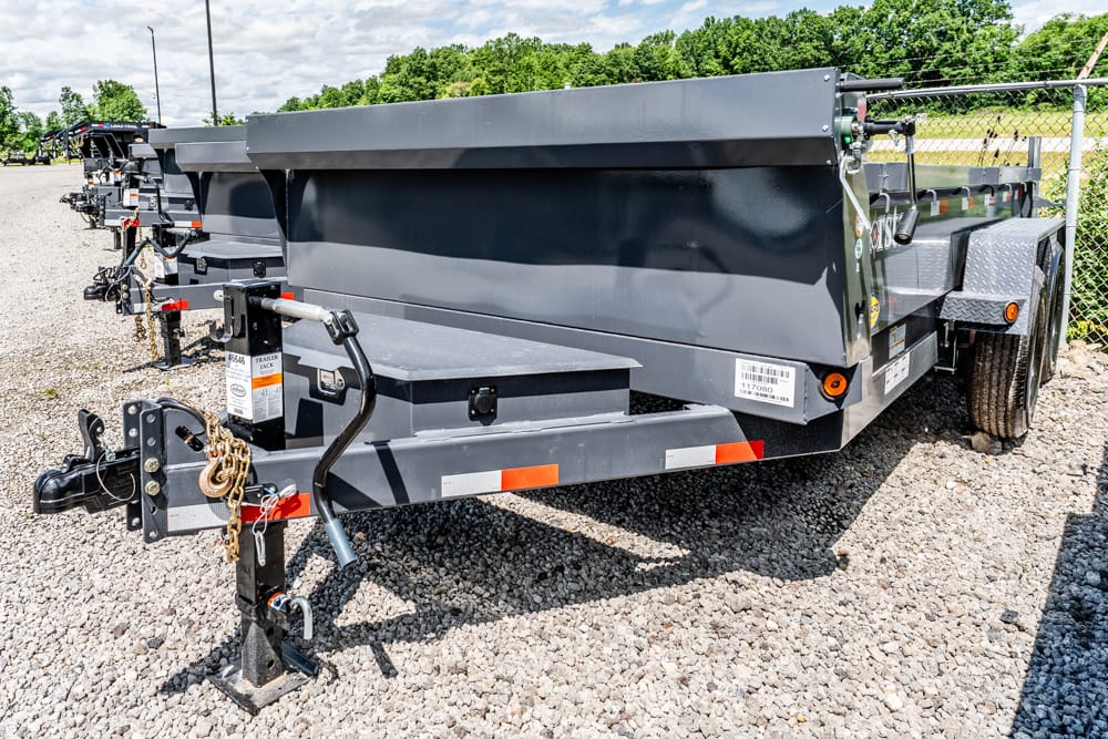 Grey utility trailer on a gravel lot, showing hitch, chains, and front jack with other trailers in the background.