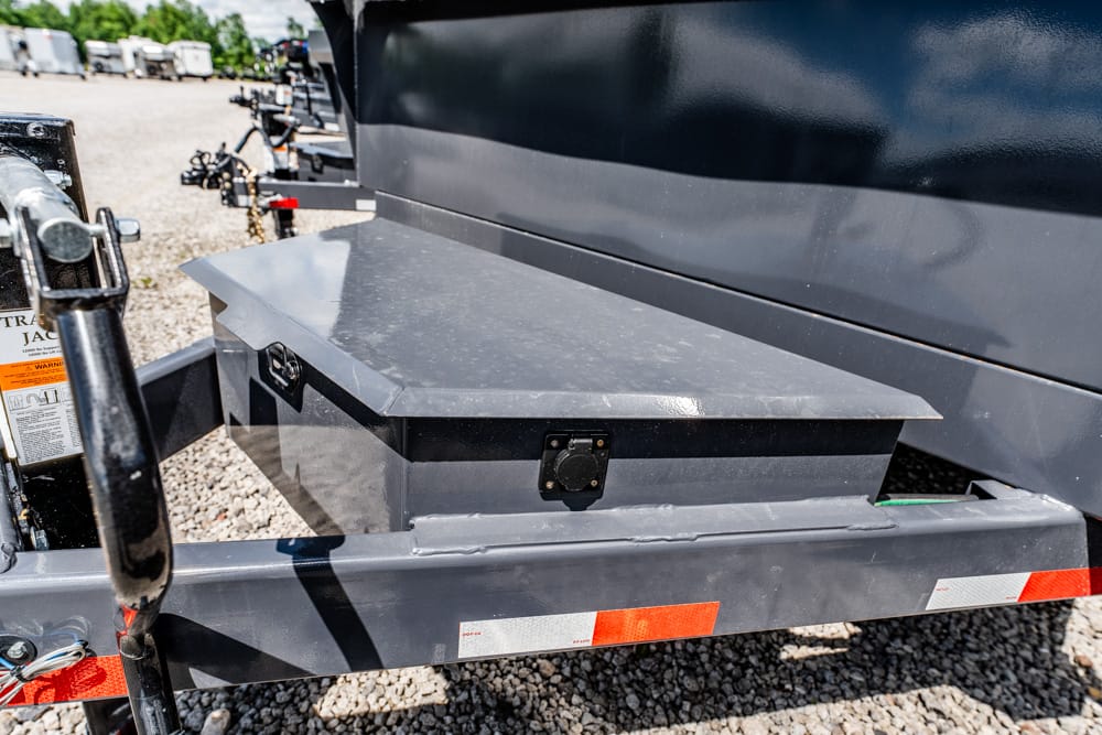 Gray metal storage toolbox mounted on a trailer frame, featuring a latch and tilt-top lid, on a gravel lot with other trailers nearby