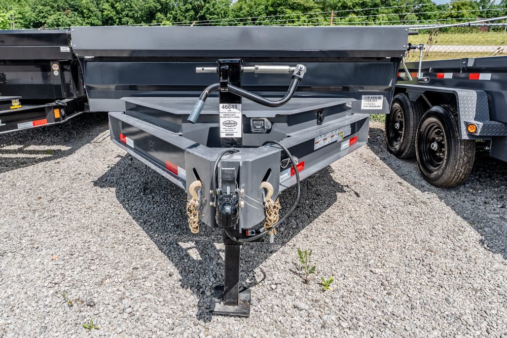 Front view of a gray utility trailer hitch with safety chains and hitch lock on a gravel lot