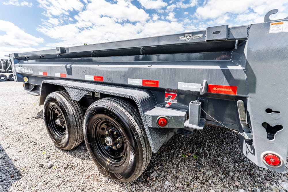Heavy-duty gray dump trailer with dual axles on a gravel lot, side rails and taillights visible.