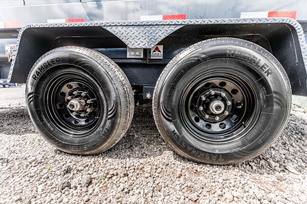 Dual-axle trailer with two black steel wheels and tires on a gravel surface under a metal fender area, labeled Rambler on the tire sidewalls