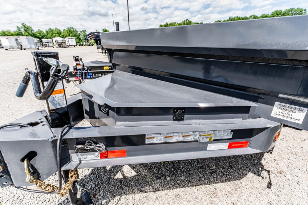 Close-up of a gray dump trailer hitch with hydraulic lift, safety chains, and control handle on a gravel lot.